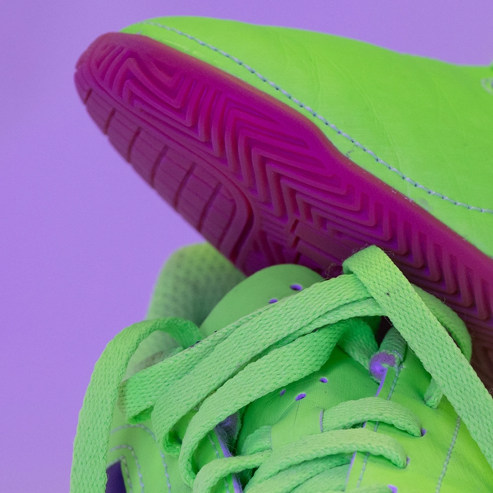 Detail eines neonfarbenen Fußballschuhs unter magentafarbenem LED-Licht mit stark veränderter Sohle und Hintergrundfarbe.