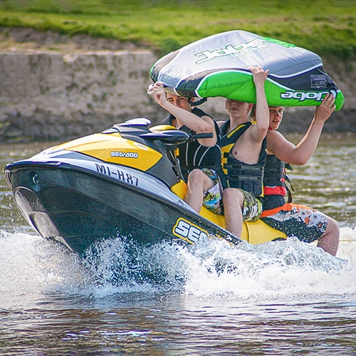 Drei Jugendliche fahren mit einem Sea-Doo Jetski über das Wasser und halten ein aufblasbares Wasserspielgerät über ihren Köpfen – actionreiche Eventaufnahme.
