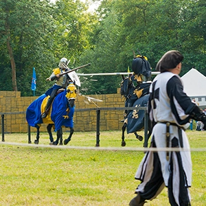 Zwei Ritter in farbigen Wappenröcken reiten beim Lanzenstechen aufeinander zu, während ein Darsteller im Vordergrund das Turnier beobachtet.
