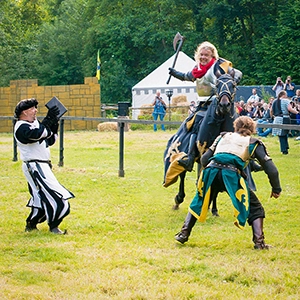 Berittener Kämpfer in mittelalterlicher Rüstung schwingt eine Waffe während einer Show auf dem MPS Bückeburg, umgeben von Darstellern und Zuschauern.