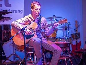 Junger Musiker sitzt auf einem Hocker und spielt Akustikgitarre, während er in ein Mikrofon singt – Bühnenlicht in Blau und Magenta.