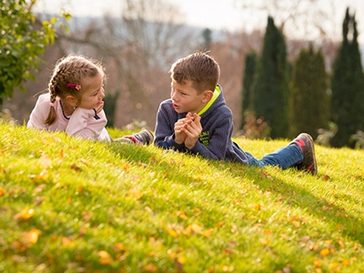 Zwei lachende Kinder, ein Junge und ein Mädchen, liegen entspannt auf einer sonnigen grünen Wiese in einem Park und unterhalten sich. Die herbstliche Stimmung und das warme Licht betonen die fröhliche Atmosphäre. Ein natürliches Familienfoto von Michael Schalansky.