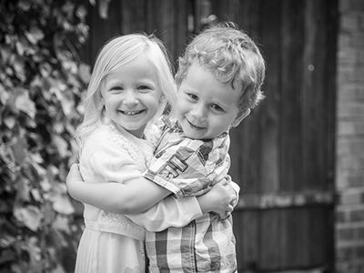 Geschwisterporträt in Schwarz-Weiß: zwei Kinder umarmen sich lachend vor Holztür und Efeu – emotionale Kinderfotografie in Erfurt.