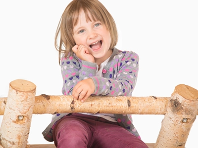 Fröhliches Mädchen-Porträt im Kindergarten: blondes Kind lacht, sitzt auf Birkenholzstamm vor hellem Hintergrund – natürliche Kinderfotografie in Erfurt.