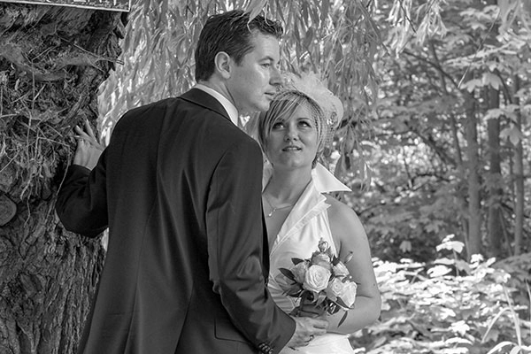 Romantisches_Brautpaar_Outdoor_Hochzeit_Thüringen Schwarz-Weiss-Hochzeitsfoto eines Brautpaares in einem natürlichen Ambiente. Der Bräutigam lehnt an einem Baum, während die Braut ihn mit einem zärtlichen Blick anschaut und ihren Brautstrauß mit weißen Rosen hält. Das Bild fängt einen intimen und romantischen Moment in der Natur ein. Hochzeitsfotografie von Michael Schalansky in Thüringen.
