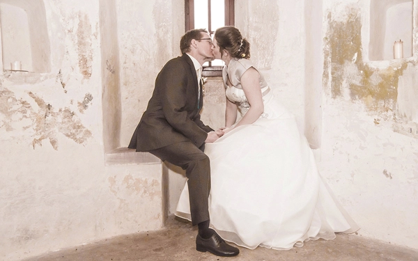 Hochzeitskuss_Historische_Location_Thüringen_Michael-Schalansky Ein romantisches Hochzeitsporträt eines Brautpaares in einer historischen Burg oder Festung in Thüringen. Das Paar sitzt eng umschlungen in einer Fensternische aus grobem Stein, küsst sich innig und hält Hände. Die Braut trägt ein weißes Kleid mit Bolero, der Bräutigam einen dunklen Anzug. Das Licht fällt sanft durch das Fenster, beleuchtet kleine Nischen mit Kerzen. Intime Hochzeitsfotografie von Michael Schalansky.