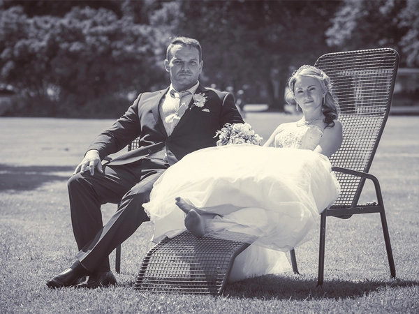 Entspanntes_Brautpaar_auf_Wiese_Hochzeit_Outdoor Schwarz-Weiss-Hochzeitsfoto eines entspannten Brautpaares in modernen Liegestühlen auf einer grünen Wiese. Der Bräutigam blickt in die Kamera, während die Braut lächelnd zur Seite schaut und die Beine ausgestreckt hat. Ein natürliches und unbeschwertes Porträt in einem Parkambiente. Beispiel für authentische Hochzeitsfotografie von Michael Schalansky.