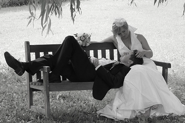 Intimer_Moment_Brautpaar_auf_Bank_Hochzeit_Natur Schwarz-Weiss-Hochzeitsfoto eines Brautpaares, das entspannt auf einer Holzbank im Freien liegt und sitzt. Der Bräutigam liegt mit dem Kopf im Schoß der Braut, die ihn liebevoll ansieht. Das Bild fängt einen zärtlichen, ungestellten Moment der Zweisamkeit in einem Park oder Garten ein. Eine authentische Hochzeitsreportage von Michael Schalansky.