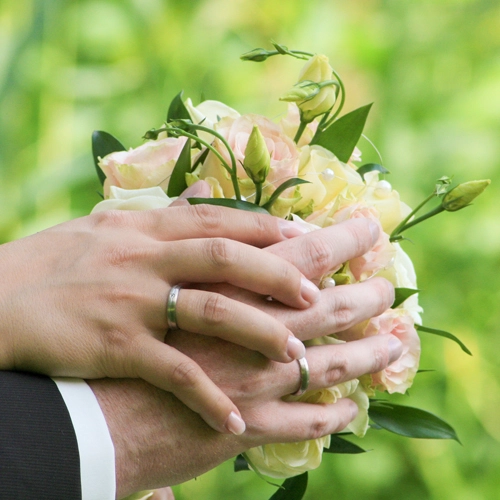 Eheringe_Brautstrauss_Hochzeit_Liebe_Symbole Nahaufnahme der Hände eines Hochzeitspaares, die gemeinsam einen Brautstrauß aus hellrosa und cremefarbenen Rosen halten. Beide tragen silberne Eheringe. Die Hand der Braut liegt sanft über der des Bräutigams. Der Hintergrund ist unscharf grün. Das Bild symbolisiert Verbundenheit und ist ein Beispiel für emotionale Hochzeitsdetailfotografie von Michael Schalansky.