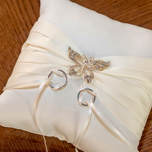 Hochzeitsringe_Kissen_Symbol_Liebe_Detailaufnahme Nahaufnahme von zwei silbernen Eheringen, die mit weißen Bändern auf einem eleganten, cremefarbenen Ringkissen liegen. Das Kissen ist mit einer glitzernden Schmetterlingsbrosche verziert. Der Hintergrund ist ein unscharfer Holztisch. Das Bild symbolisiert die Liebe und Verbundenheit und ist ein Beispiel für detaillierte Hochzeitsfotografie von Michael Schalansky.