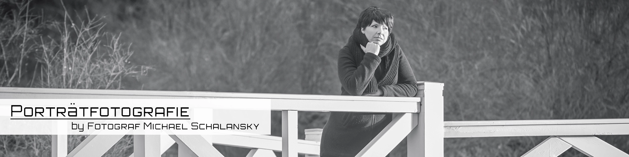 Ausdrucksstarkes Schwarz-Weiß-Portrait einer Frau auf einer Brücke. Ihre rechte Hand stützt das Kinn, während sie mit ernstem Ausdruck zur Seite blickt. Sie trägt dunkle Winterkleidung. Das weiße Brückengeländer bildet einen Kontrast zum dunklen, unscharfen Hintergrund aus Vegetation. Aufgenommen von Fotograf Michael Schalansky.