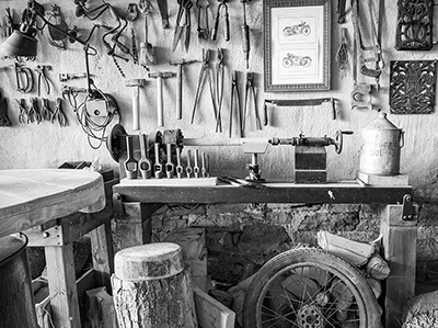 Nahaufnahme einer Wand voller verschiedener, teils verrosteter Metallwerkzeuge mit hölzernen Griffen, die ihre individuelle Textur und die Spuren der Nutzung zeigen, darunter eine rustikale Werkbank, alles in Schwarz-Weiß, detailreiche Aufnahme von Michael Schalansky.