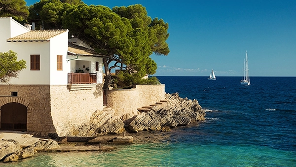 Eine Komposition aus einem weißen Haus, grünen Bäumen, braunen Felsen und dem tiefblauen bis türkisfarbenen Meer, beleuchtet von der Sonne, das die Farbpalette der mediterranen Küste zeigt, Fotografie von Michael Schalansky.