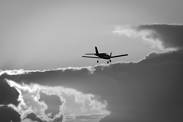 Ein kleines Propellerflugzeug im Flug, als Silhouette vor einem dramatischen, wolkigen Himmel in Schwarz-Weiß, symbolisch für Freiheit und Reisen, Luftfahrtfotografie von Michael Schalansky.