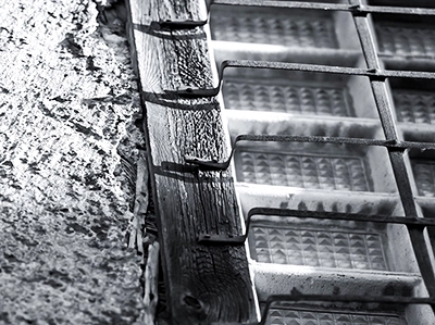 Detailansicht einer alten, verwitterten Wand mit rauer Oberfläche, einem vertikalen Holzbalken und einem Fenster aus strukturierten Glasbausteinen mit rostigem Gitter, typisch für Urban Exploration, Michael Schalansky Fotografie.