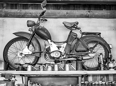 Ein klassisches Mofa mit Speichenrädern und Ledersattel steht auf einem Werkstattregal, umgeben von verschiedenen Werkzeugen und einer Lampe an einer strukturierten Wand, alles in Schwarz-Weiß, Industriefotografie von Michael Schalansky.