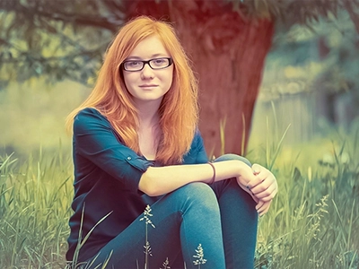 Jugendporträt einer jungen Frau mit roten Haaren und Brille, sitzend im Gras unter einem Baum.