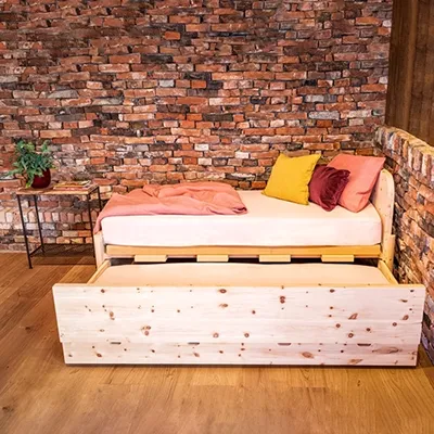 Produktbild Bett Funktionsaufnahme mit Zubehör Detailaufnahme einer warm und gemütlich eingerichteten Studioecke: Ein helles Holzbett mit herausziehbarer Liegefläche, dekoriert mit rosafarbenen und gelben Kissen, steht vor einer markanten roten Backsteinwand. Die Szene strahlt eine behagliche Atmosphäre aus.