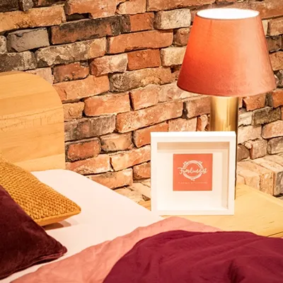 Detailansicht Fotostudio – Warme Einrichtung für Shootings Nahaufnahme eines Bettenbereichs in einem Fotostudio mit Backsteinwand, warmen Rottönen bei der Bettwäsche und einer stimmungsvollen Lampe. Ein Markenlogo "Familiennest – Fotografie mit Herz" ist auf einem Rahmen zu sehen, was auf ein Studio für Familienfotografie hindeutet.