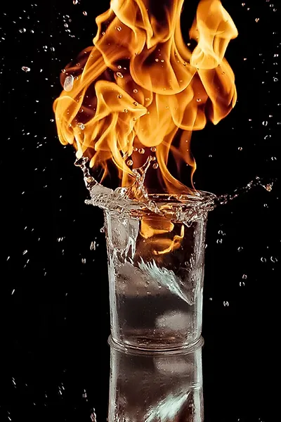 Visuelle Emotion: Feuer und Wasser im Glas. Beispiel für kreative und ausdrucksstarke Produktfotografie von Michael Schalansky. Kreative Produktfotografie: Glas mit Eiswürfeln, aus dem Flammen mit Wasserspritzern aufsteigen, vor schwarzem Hintergrund. Fotografiert von Michael Schalansky.