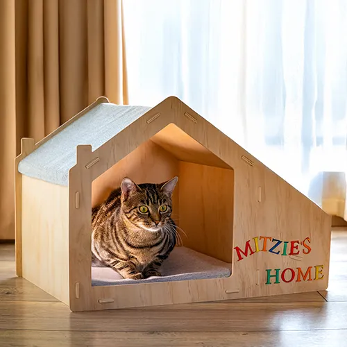 Getigerte Katze liegt im Holz-Katzenhaus mit personalisiertem Schriftzug „MITZIE’S HOME“ im warmen Wohnraumlicht.
