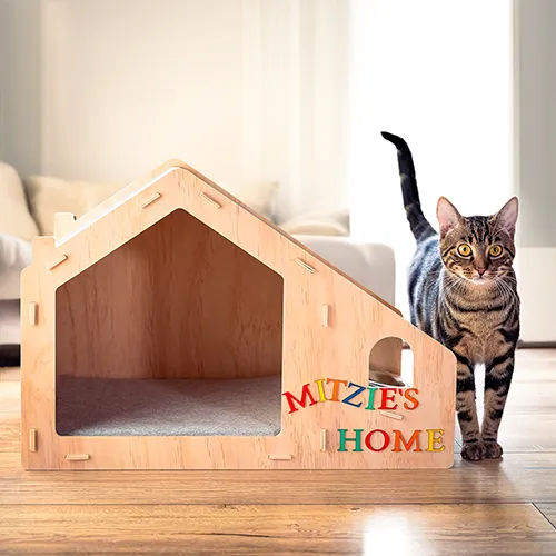 Holz-Katzenhaus mit personalisiertem Schriftzug „MITZIE’S HOME“ im Wohnraum, daneben steht eine getigerte Katze mit erhobenem Schwanz.