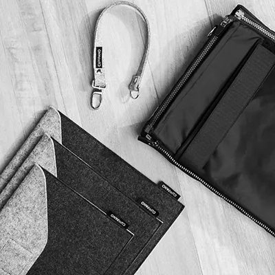 Accessoire-Fotografie: Cumpano Organizer und Handschlaufe. Präzise Produktbilder für Onlineshops und Modekataloge. Zubehör-Set von Cumpano: drei Filz-Organizer in Dunkelgrau und eine helle Handschlaufe mit Karabiner, auf hellem Holzboden. Detailreiche Produktfotografie.