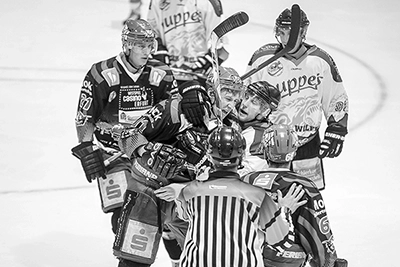Ein Schwarz-Weiß-Foto, das den Moment nach einem Zweikampf im Eishockey zeigt, bei dem sich Spieler beider Teams um den Schiedsrichter versammeln. Die Gesichter der Spieler zeigen Anspannung und Konzentration. Die Trikots der Black Dragons sind dunkel, die der Gegner hell.