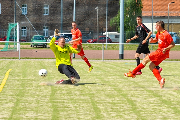 Ein Torwart des FC Union Erfurt im grünen Trikot ist im Hechtsprung zu sehen, um einen niedrig rollenden Fußball abzuwehren, während ein gegnerischer Spieler im roten Trikot einen Schuss abgibt. Die Szene spielt auf einem Kunstrasenplatz.